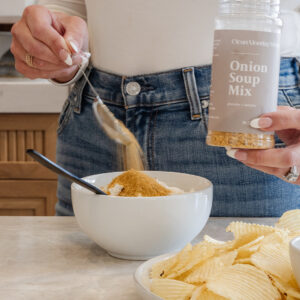 A woman holding Clean Monday Meals Onion Soup Mix, sprinkles some into a dip bowl with a platter of chips in the foreground.
