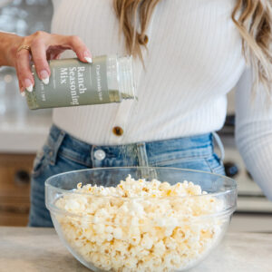 A woman sprinkles Clean Monday Meals ranch seasoning mix onto a bowl of popcorn in the kitchen.